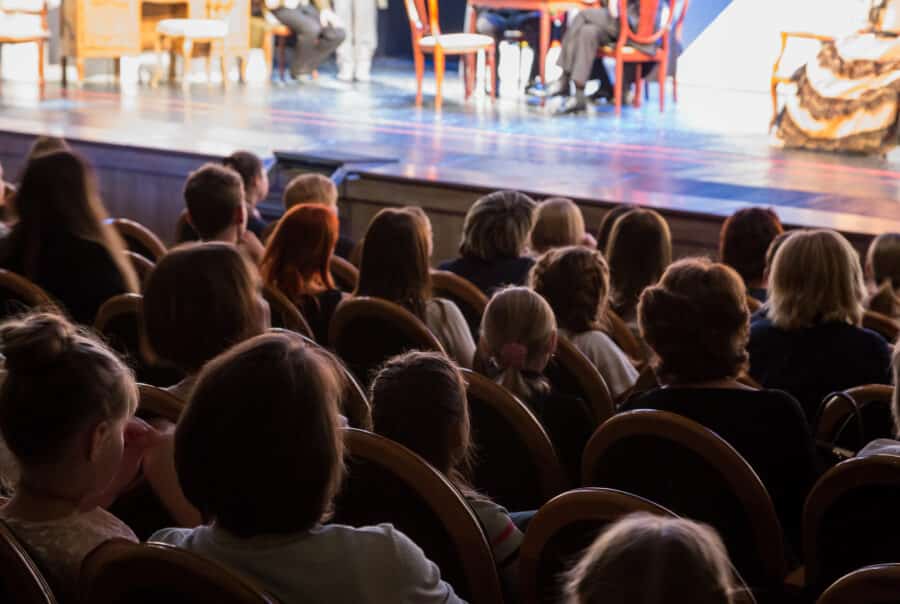 Crowd of audience members seated in a theater, facing a brightly lit stage.
