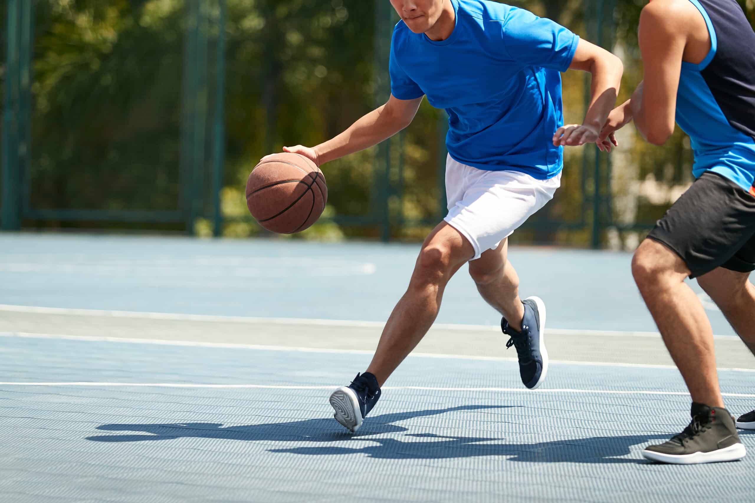 Two players in blue shirts chase a basketball on an outdoor court, their legs in motion.