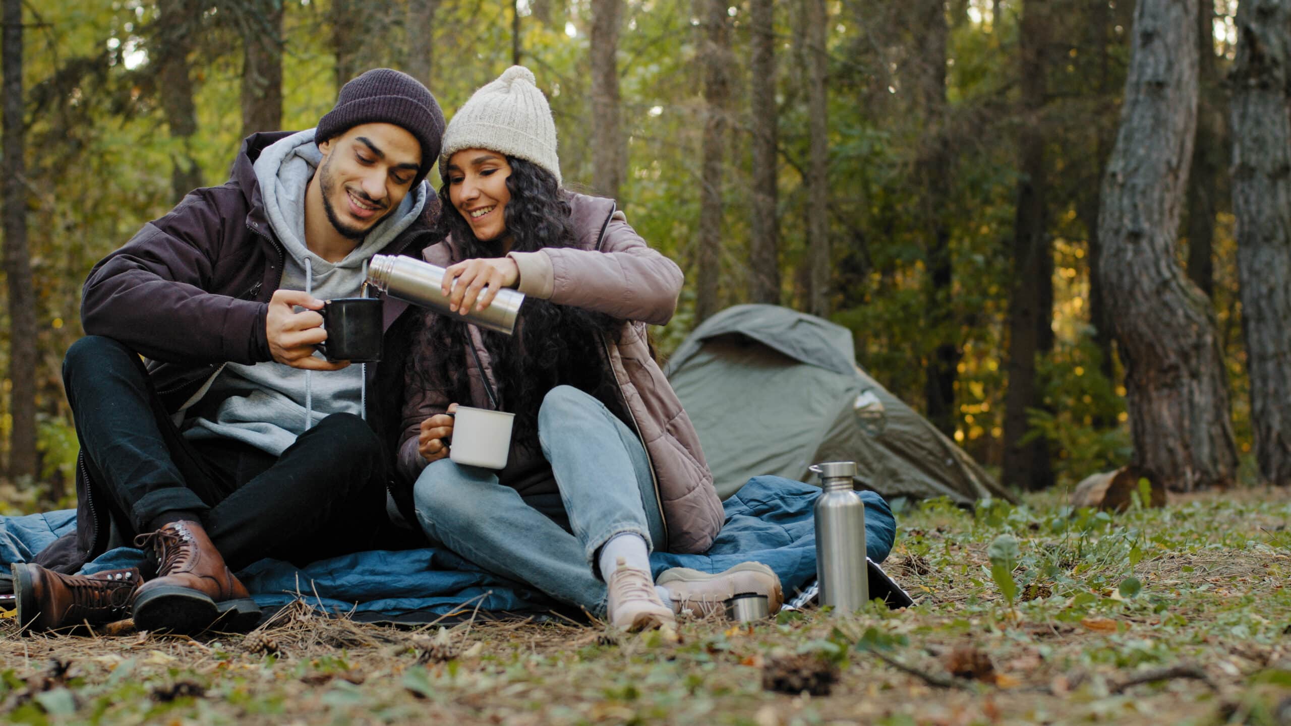 Two friends sit on a tarp in a forest campsite as one pours a drink from a thermos into a mug, smiling at each other with a tent in the background.